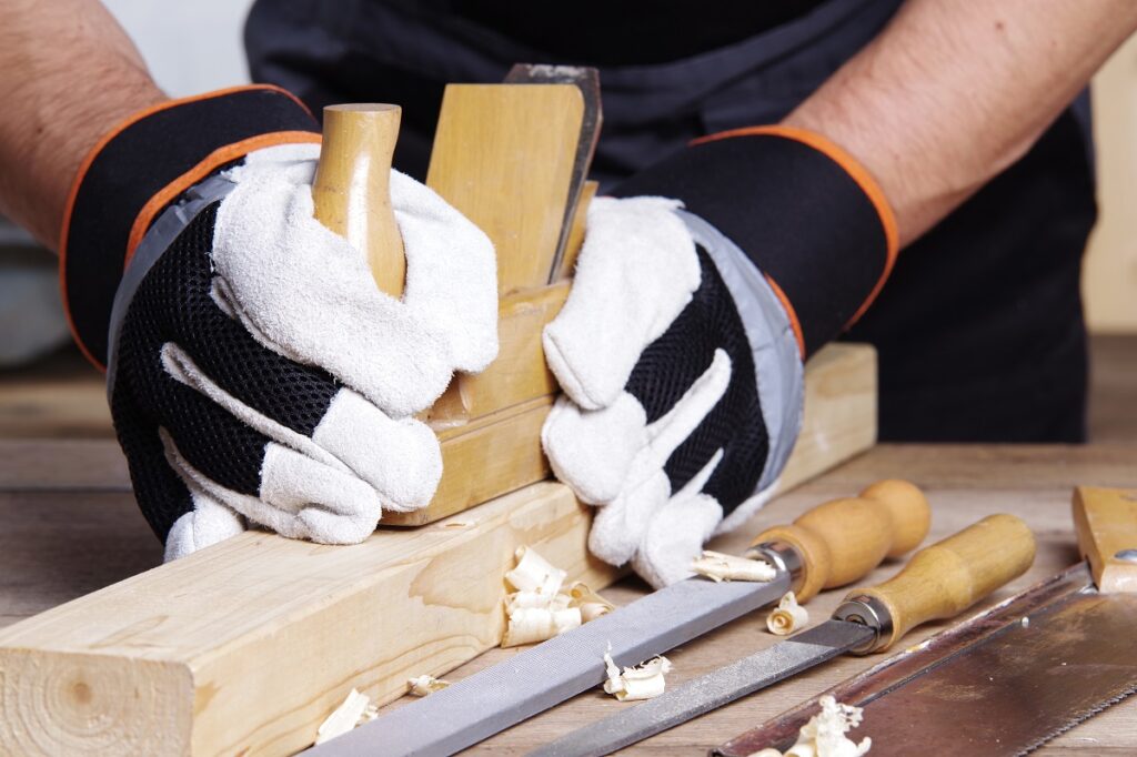 Ein Schreiner hobelt mit weiß-schwarz-orangen Arbeitshandschuhen aus Leder und Nylon ein Stück Holz.
