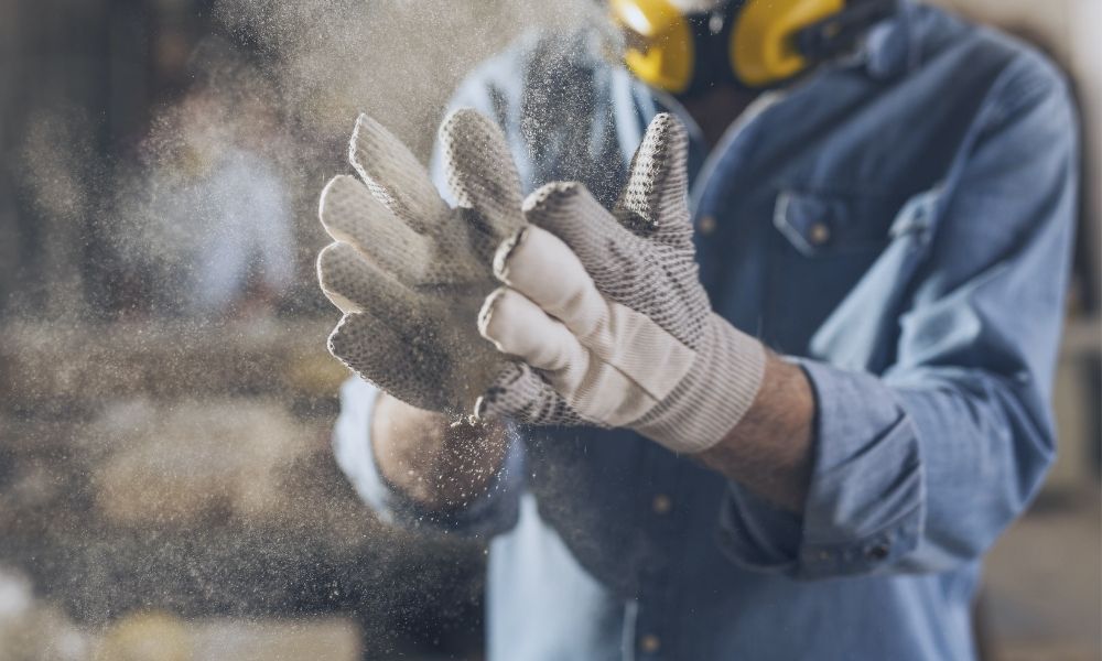 Ein Schreiner mit Gehörschutz klopft Sägemehl von seinen Baumwoll-Handschuhen. Die Arbeitshandschuhe sind mit Gummi-Noppen versehen.