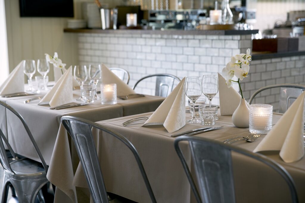 2 Tische in einem Restaurant sind elegant eingedeckt mit grauer Duni-Tischdecke und weißen, struckturierten Duni-Servietten.